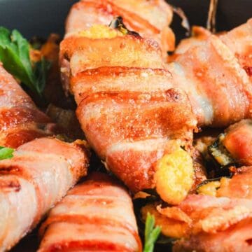 Bacon-wrapped jalapeño poppers are arranged on a plate. The poppers appear golden and crispy, with visible cheese oozing out. Fresh parsley leaves are used as garnish.