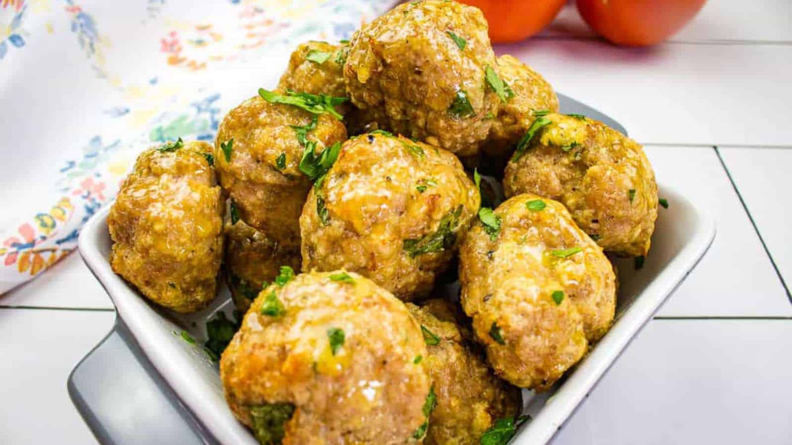 A white square dish filled with golden-brown baked meatballs garnished with fresh parsley, perfect for quick dinner solutions. The dish is placed on a white surface with a floral cloth and two tomatoes in the background.