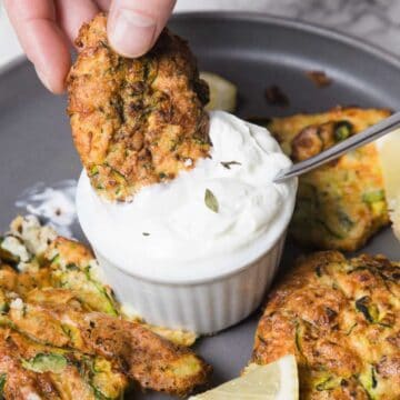 A hand dipping a zucchini fritter into a small dish of white dipping sauce on a gray plate. The plate also contains additional fritters and lemon wedges. A spoon is in the dish, and a piece of lemon is visible on the marble surface nearby.