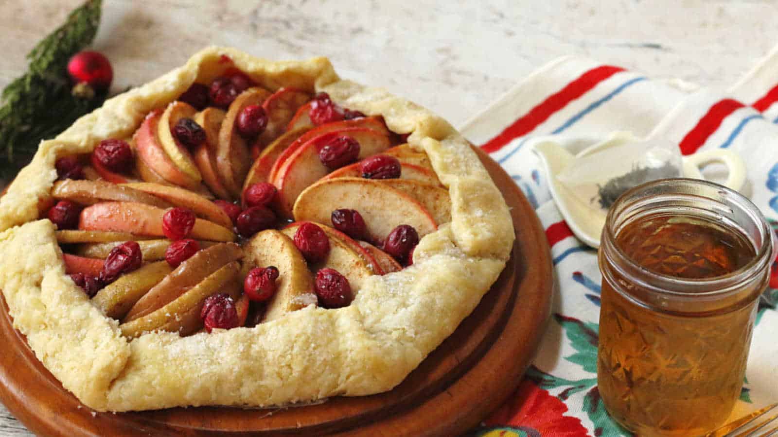 A rustic apple and cranberry galette is displayed on a wooden board. The pie has a golden, flaky crust, and is accompanied by a jar of honey. A colorful cloth and a small white dish are in the background.