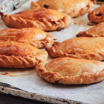 A baking tray with seven golden-brown pastry turnovers, likely empanadas, on a sheet of parchment paper. The pastries appear freshly baked, with slightly crispy edges, resting on a rustic, worn metal tray.