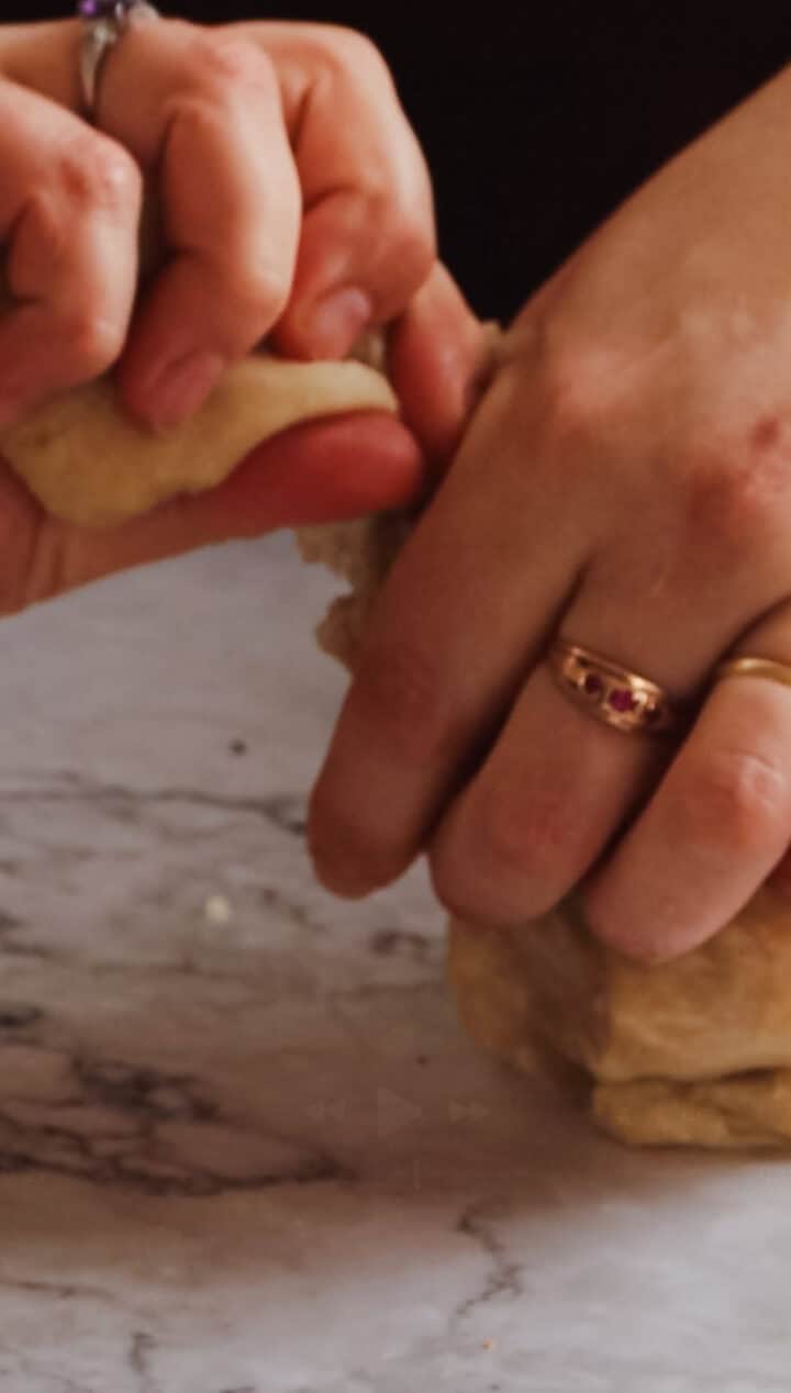 Close-up of hands kneading Argentinian empanada dough on a marble surface. One hand presses the dough while the other holds it gently. The person wears rings, including a notable one with a red gemstone.