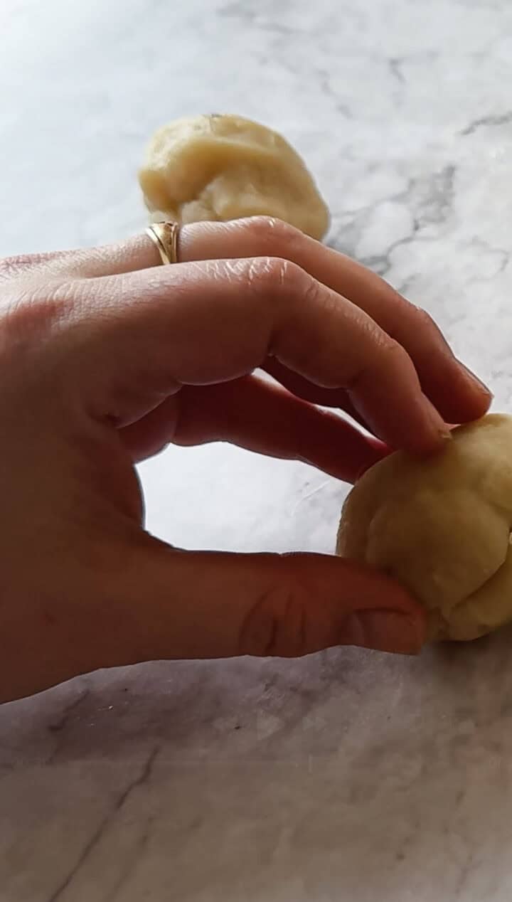 A hand with a ring is delicately shaping a ball of Argentinian empanada dough on a marble countertop. Another ball of dough is visible in the background, waiting to be transformed into delicious parcels.