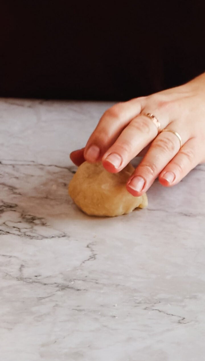 Close-up of a hand with painted nails and a ring, pressing on a ball of Argentinian empanada dough on a marble countertop. The dough appears to be in the process of being shaped or kneaded.