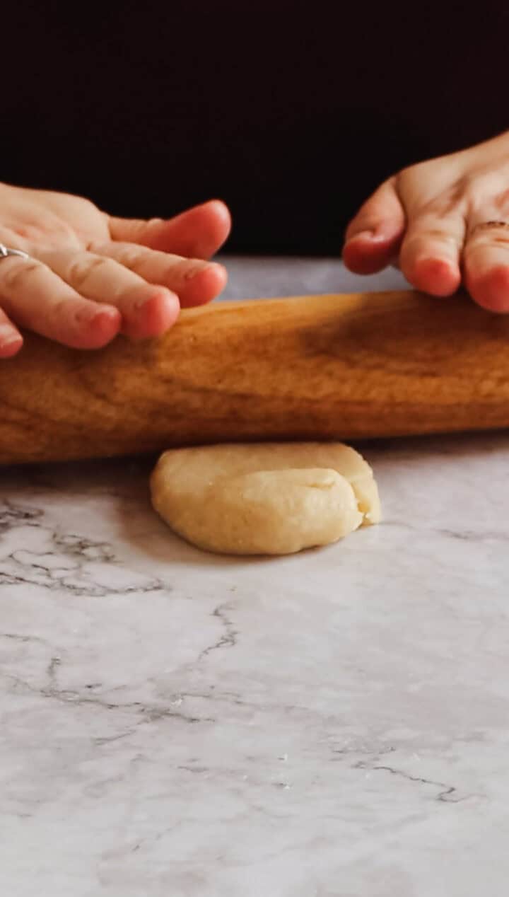 Hands with painted nails are using a wooden rolling pin to flatten a small, round piece of Argentinian empanada dough on a marbled surface.