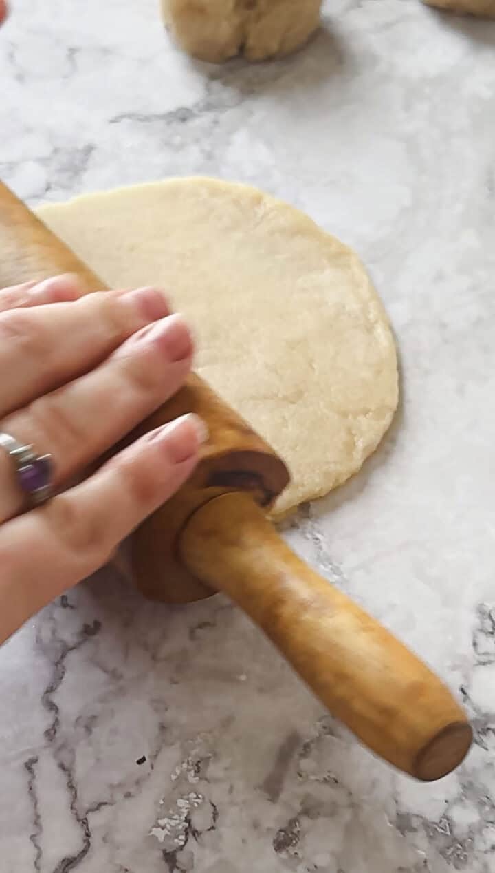 A hand with a ring is using a wooden rolling pin to flatten Argentinian empanada dough on a marble countertop. Another piece of the delicate dough is visible in the background.