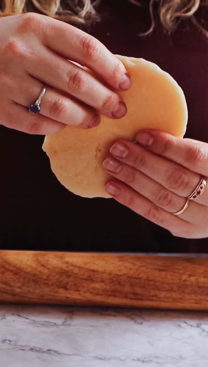 A person is expertly holding a circular piece of Argentinian empanada dough with both hands, their fingers adorned with rings. Below, a wooden surface adds rustic charm to the scene.