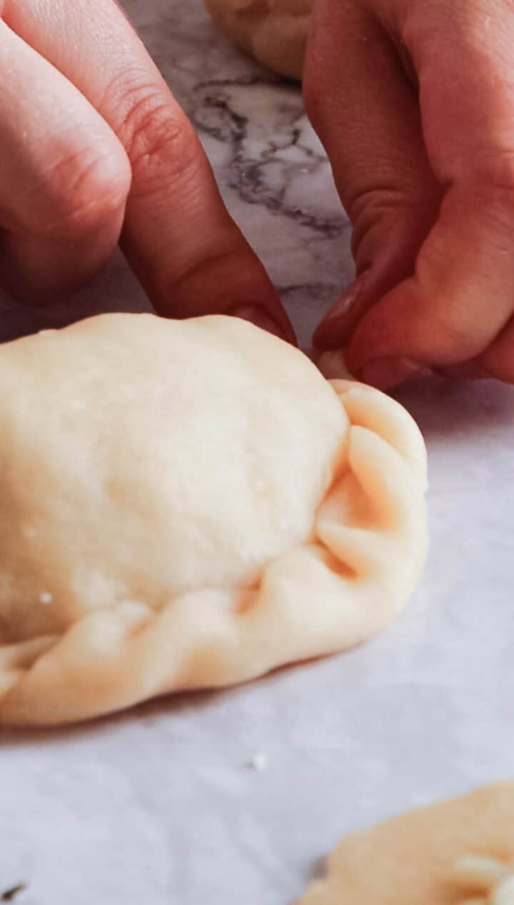 Close-up of hands pinching the edges of Argentinian empanada dough to seal it. The dough rests on a white marble surface as the hands expertly crimp and pleat its edge.