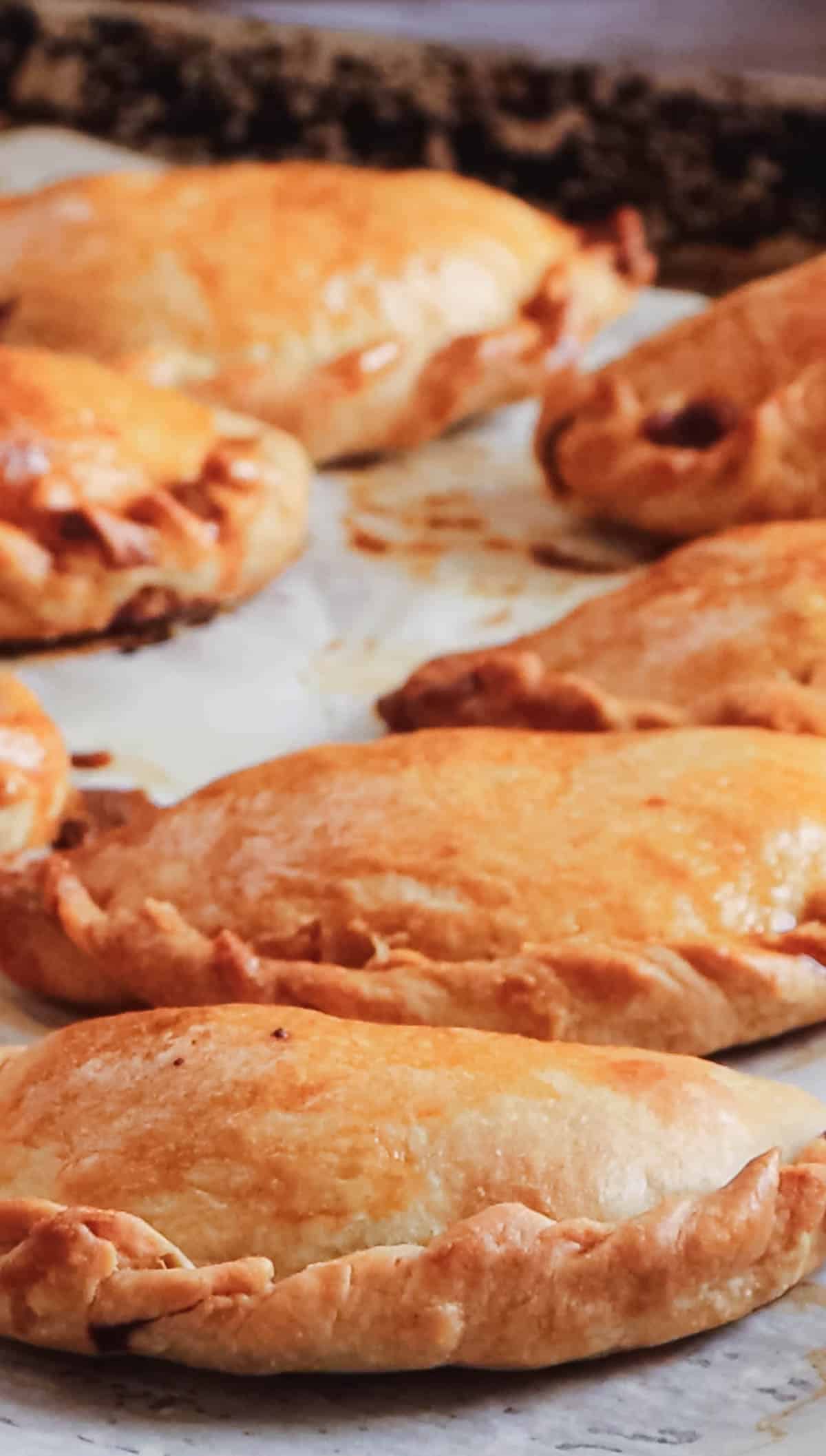 A tray of freshly baked Argentinian empanadas rests on parchment paper, their golden-brown crusts shaped into perfect half-moons. The pastries, made with traditional empanada dough, glisten with an egg wash as tantalizing fillings peek out temptingly.