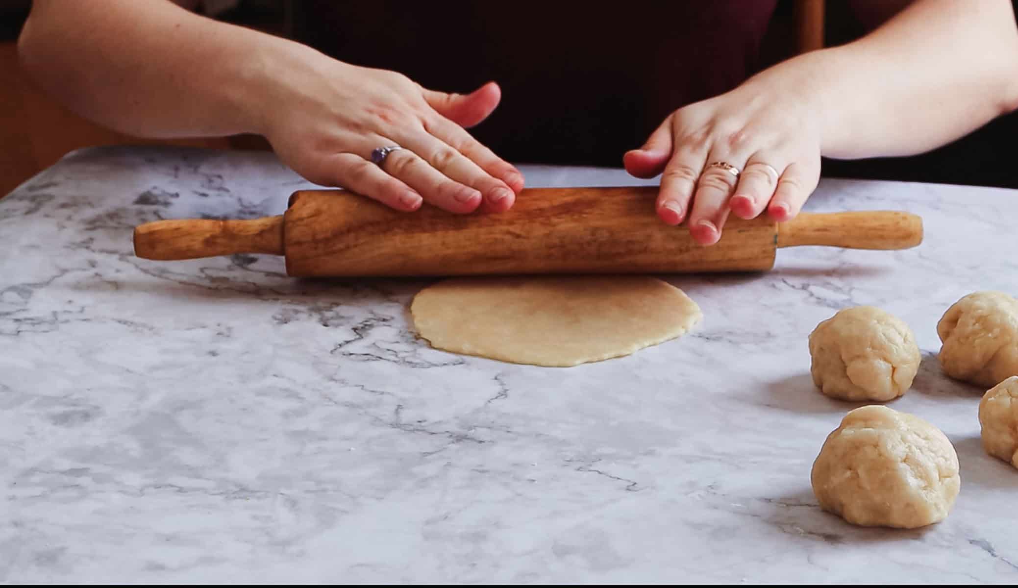 A person skillfully rolls out Argentinian empanada dough with a wooden rolling pin on a cool marble surface. Two dough balls rest on the right side, ready to be shaped into delicious treats by their expert hands.