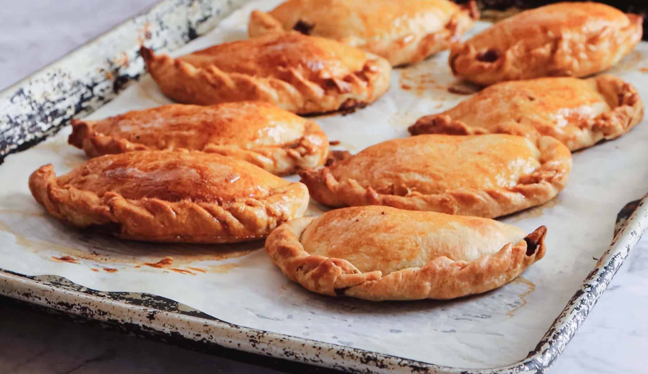 Eight golden-brown pastries are placed on a parchment-lined baking sheet. The sheet has a worn, rustic appearance, and the pastries are arranged in two rows. They have a braided edge and appear freshly baked.