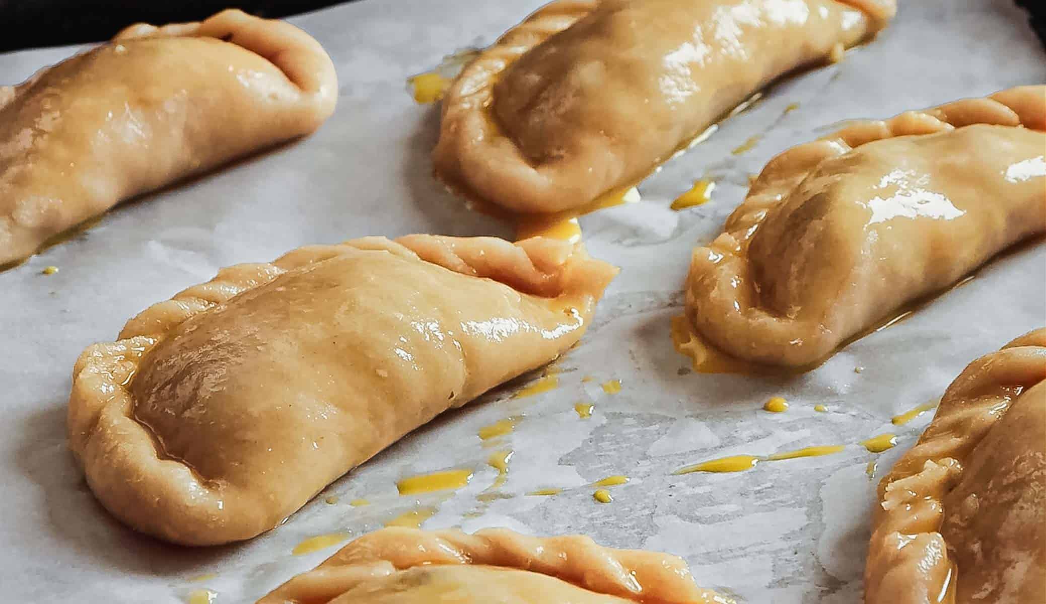 Six uncooked Argentinian empanadas arranged on a baking sheet lined with parchment paper. The pastries have a golden sheen, likely from an egg wash, and show crimped edges that seal the Argentinian empanada dough. Some filling appears to be seeping slightly from a few.