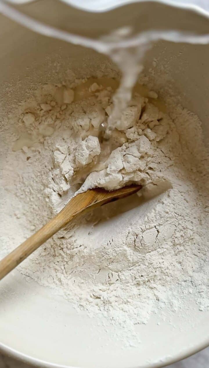 Water is being gently poured into a bowl containing flour, signaling the start of crafting Argentinian empanada dough. A wooden spoon rests inside, partially buried in the flour, ready to blend these simple ingredients into a delicious creation.