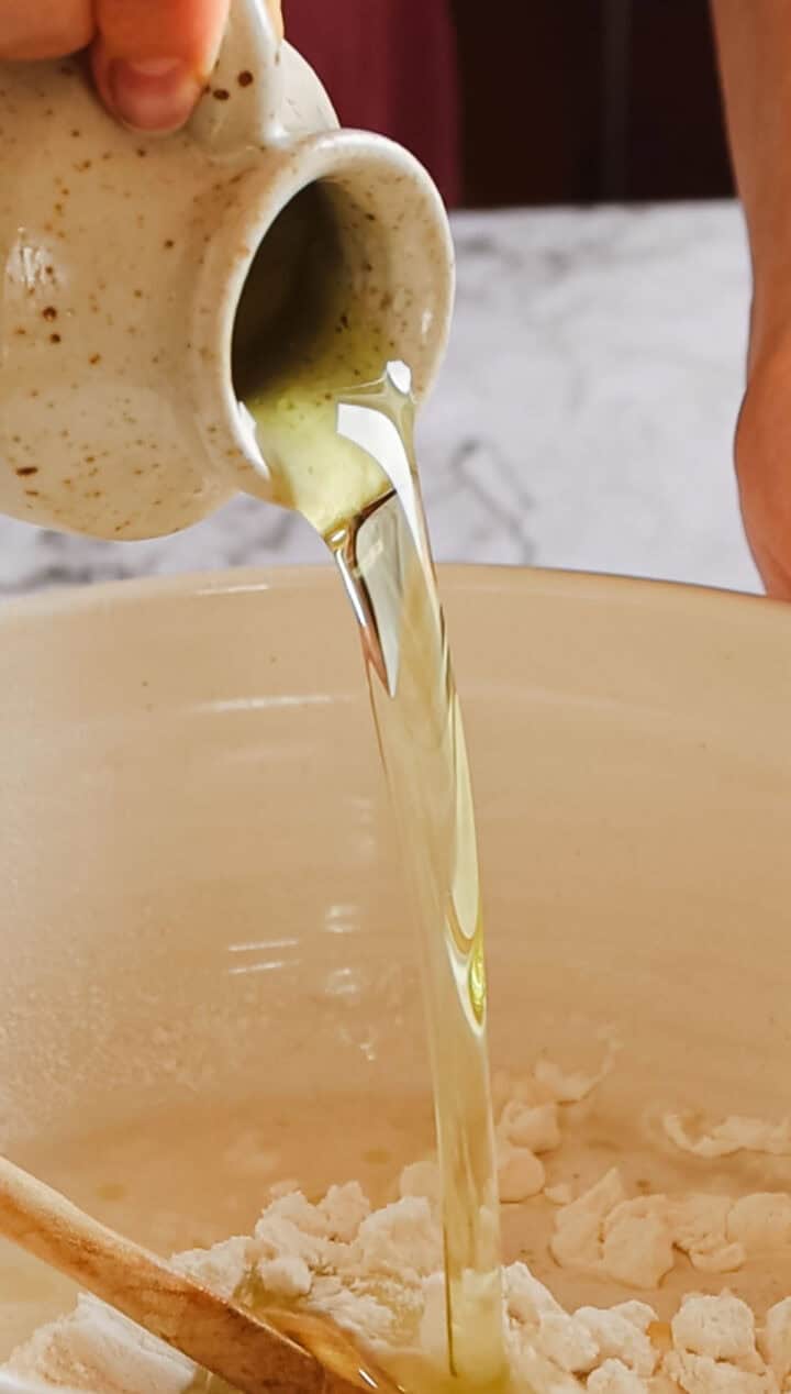 A hand pours golden-yellow liquid into a large, off-white mixing bowl filled with white flour, perfectly blending the ingredients for Argentinian empanada dough. The blurred background hints at a marble surface, enhancing the serene kitchen scene.