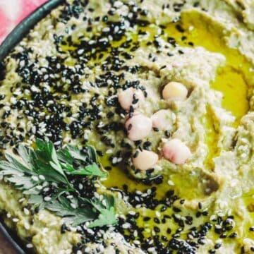A bowl of green hummus garnished with black sesame seeds, whole chickpeas, and a drizzle of olive oil. There is a sprig of parsley on top. Sliced pink watermelon radishes and pita bread are on the side.