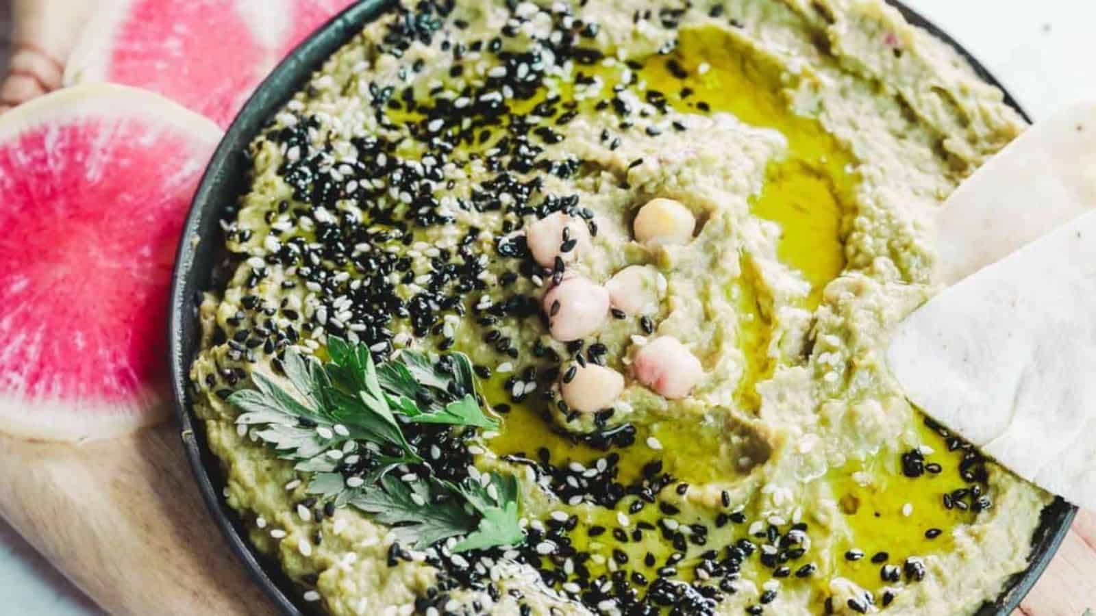 A bowl of hummus garnished with black sesame seeds, chickpeas, parsley, and olive oil, sits on a wooden board. Slices of pink and white watermelon radish are placed alongside.