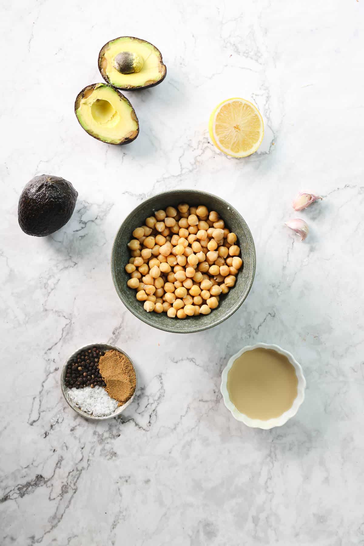 A flat lay of Avocado Hummus essentials on a marble surface, featuring a bowl of chickpeas, two halved avocados, a lemon half, two garlic cloves, and small bowls filled with pepper, cumin, salt, and tahini.