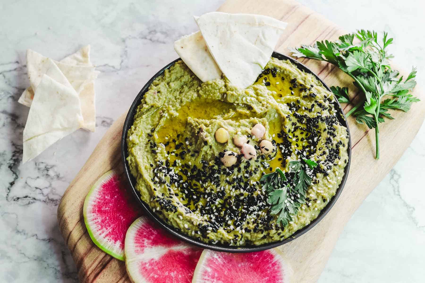 A bowl of green avocado hummus topped with chickpeas, black sesame seeds, and parsley sits on a wooden board. Pita triangles and watermelon radish slices are nearby. The avocado hummus boasts a drizzle of olive oil on top.