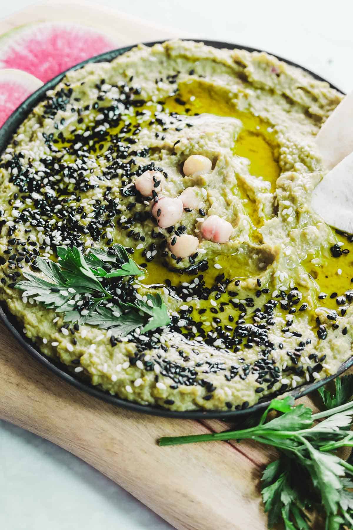 A bowl of green avocado hummus garnished with black and white sesame seeds, chickpeas, and parsley, drizzled with olive oil. The bowl rests on a wooden board, surrounded by vibrant watermelon radish slices.