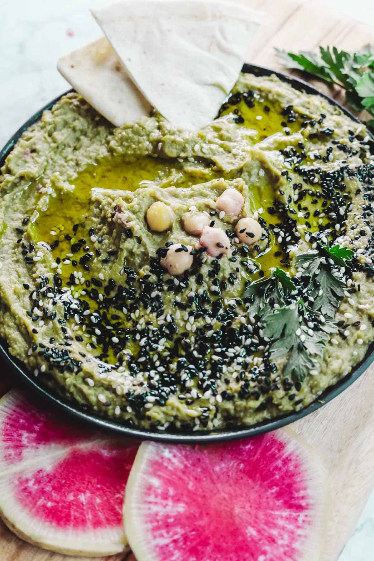 A bowl of green avocado hummus garnished with black and white sesame seeds, whole chickpeas, parsley, and olive oil. Two pita triangles rest on the side. Slices of watermelon radish are placed in the foreground on a wooden board.