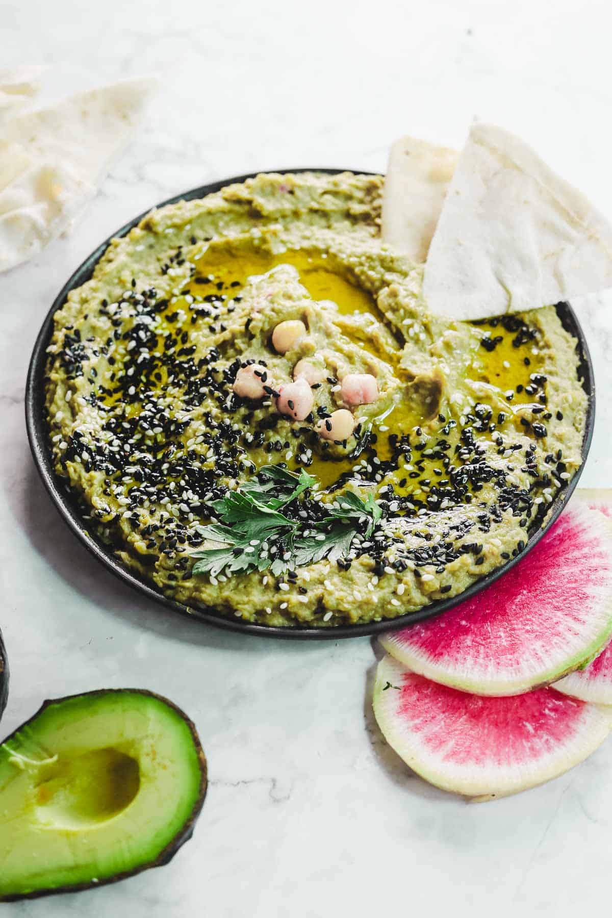 A plate of creamy avocado hummus garnished with chickpeas, parsley, and black sesame seeds, drizzled with olive oil. It's surrounded by pita triangles, sliced watermelon radish, and half an avocado on a white surface.