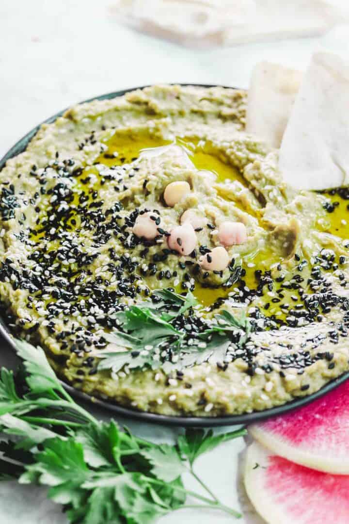 A bowl of creamy avocado hummus topped with chickpeas, black sesame seeds, and olive oil. A sprig of parsley and sliced pita bread sit invitingly beside it. In the foreground, a radish slice and more parsley garnish the light-colored surface.