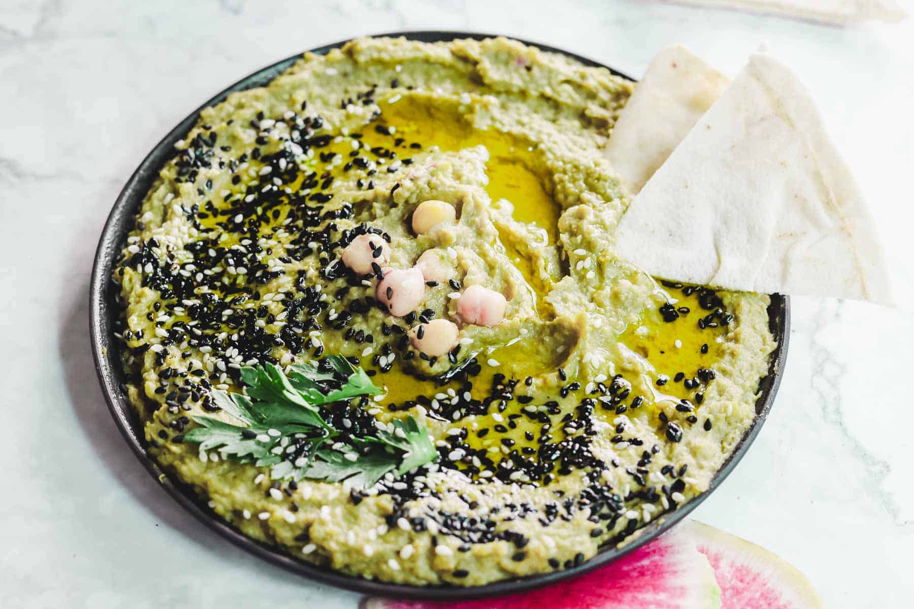 A plate of vibrant avocado hummus topped with whole chickpeas, black sesame seeds, a drizzle of olive oil, and sprigs of parsley. Two pieces of pita bread are on the side. The dish rests elegantly on a sleek marble surface.