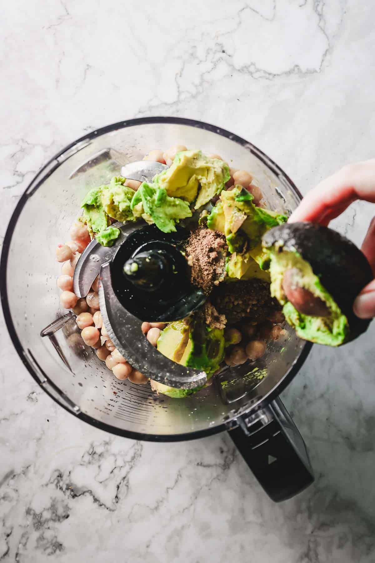 A food processor on a marble countertop contains chickpeas, avocado, and spices—a perfect blend for creamy Avocado Hummus. A hand is adding half an avocado.