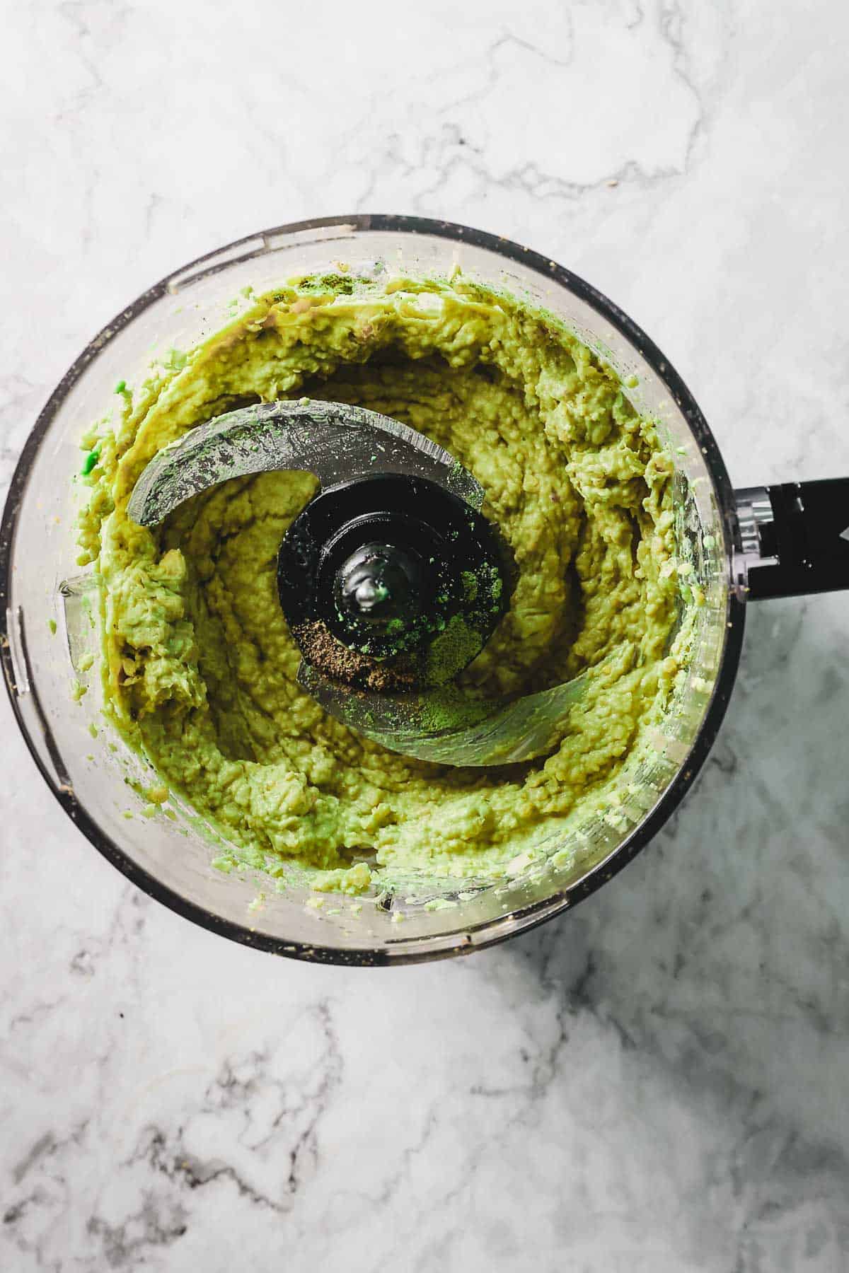 A food processor filled with a blended green avocado hummus mixture sits on a marble countertop. The visible blades are surrounded by the chunky blend, hinting at the preparation of guacamole or a creamy avocado spread.