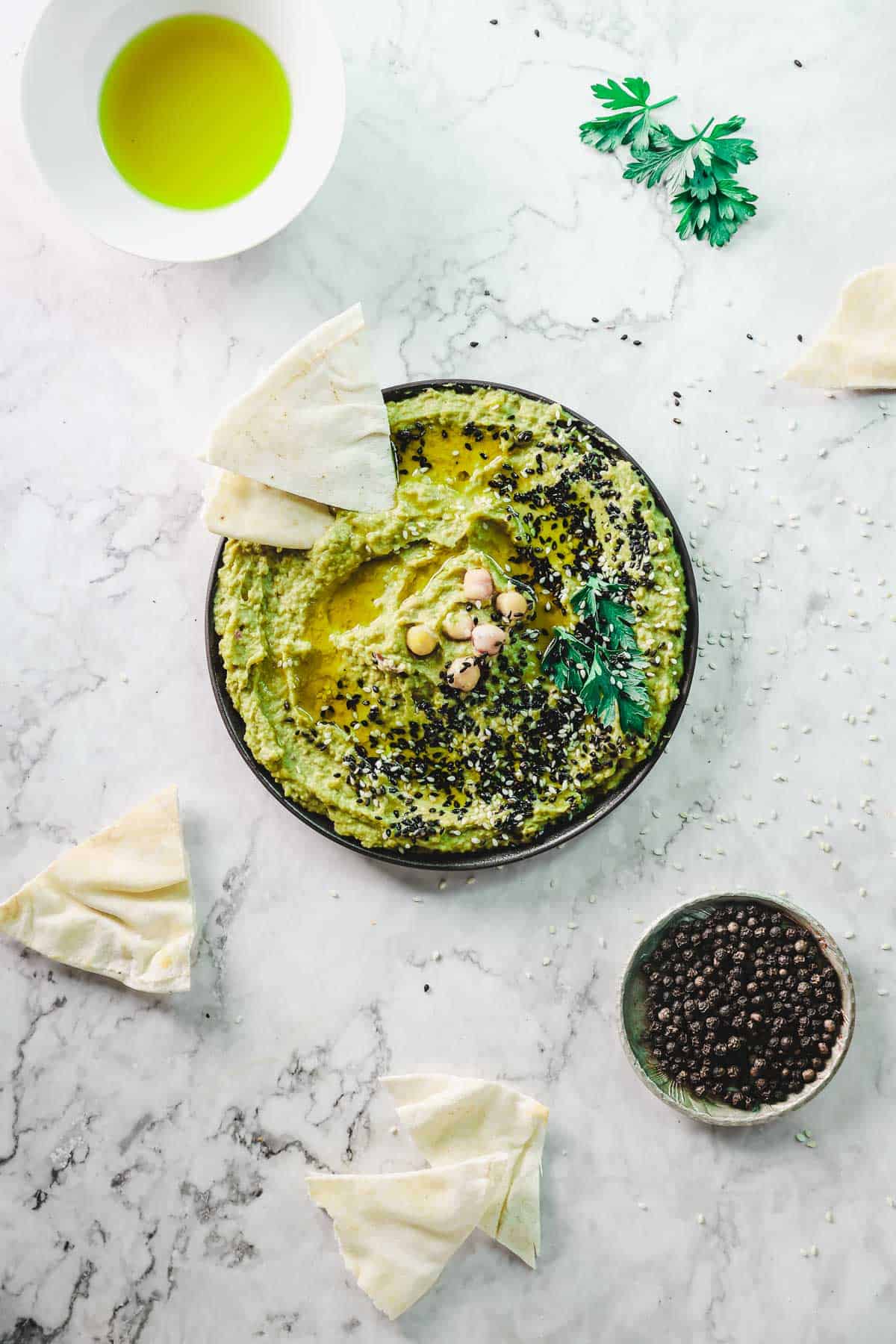 A plate of creamy avocado hummus topped with chickpeas, black sesame seeds, and parsley is surrounded by triangular pieces of pita bread. A small bowl of black peppercorns and a cup of olive oil are nearby on a marble surface.