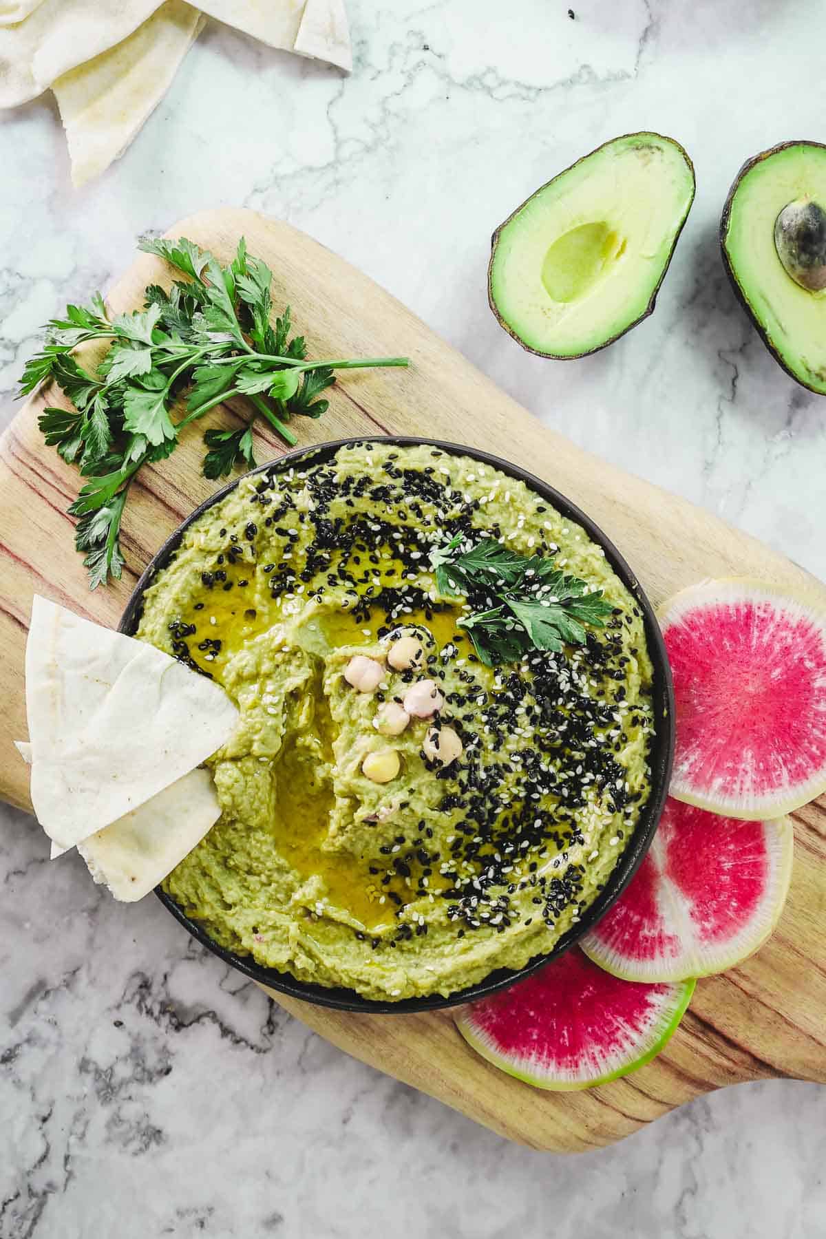 A bowl of creamy avocado hummus is topped with sesame seeds and olive oil, resting on a wooden board. Nearby, slices of watermelon radish, warm pita bread, parsley sprigs, and fresh avocado halves adorn the marble surface.