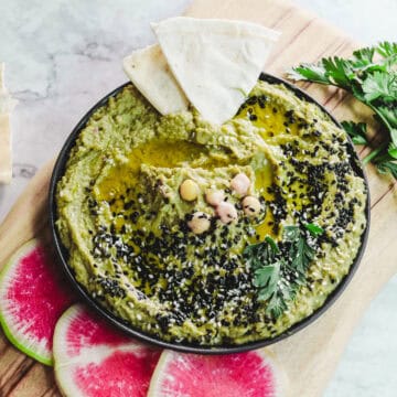 A bowl of avocado hummus topped with black sesame seeds and chickpeas, drizzled with olive oil. Garnished with parsley. Two slices of pita and watermelon radish slices are on the wooden board beside the bowl.