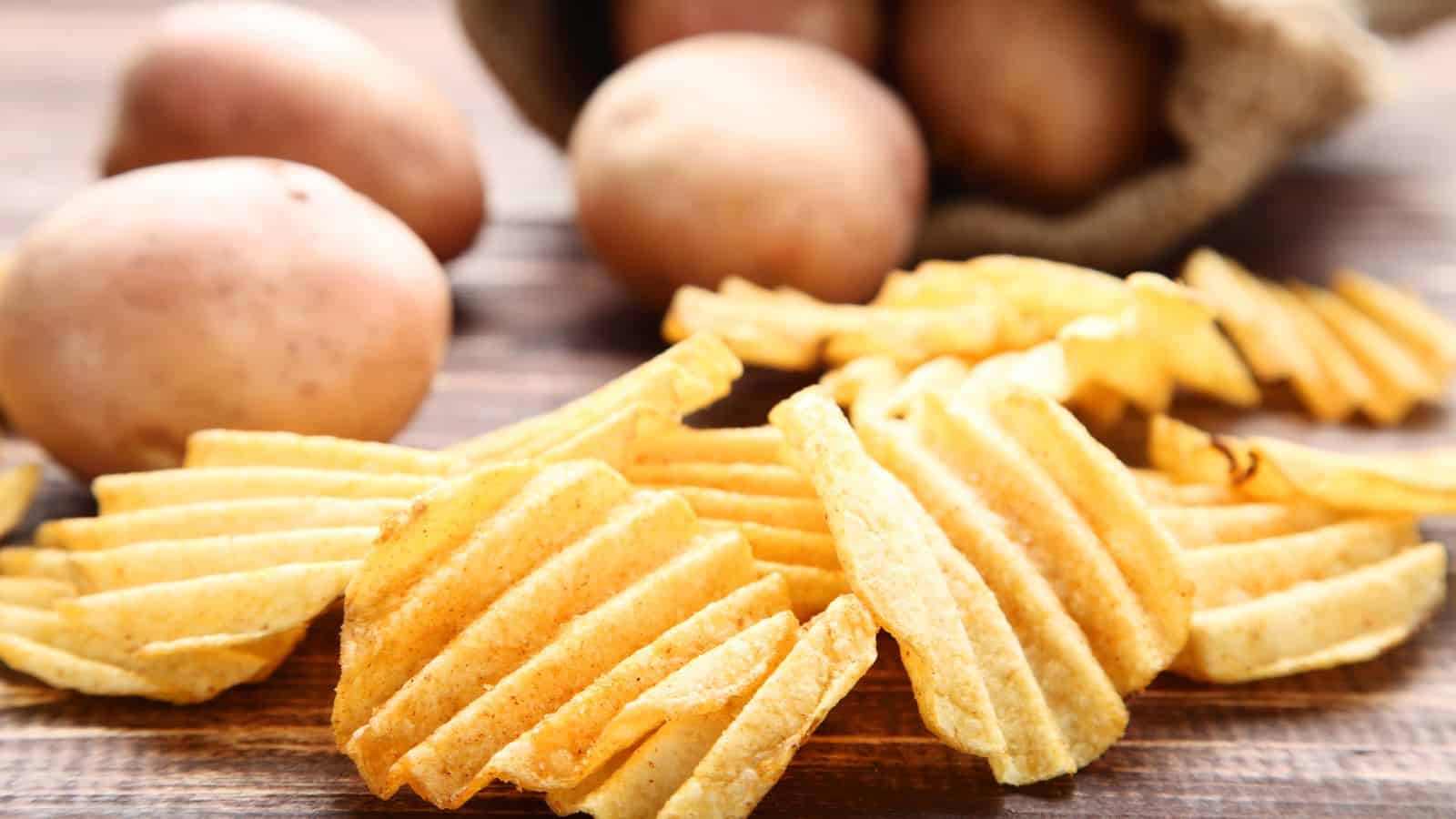 Wavy potato chips are scattered in the foreground on a wooden surface. Behind them, several whole potatoes spill out of a burlap sack. The scene suggests a natural setting for displaying the chips and raw potatoes.