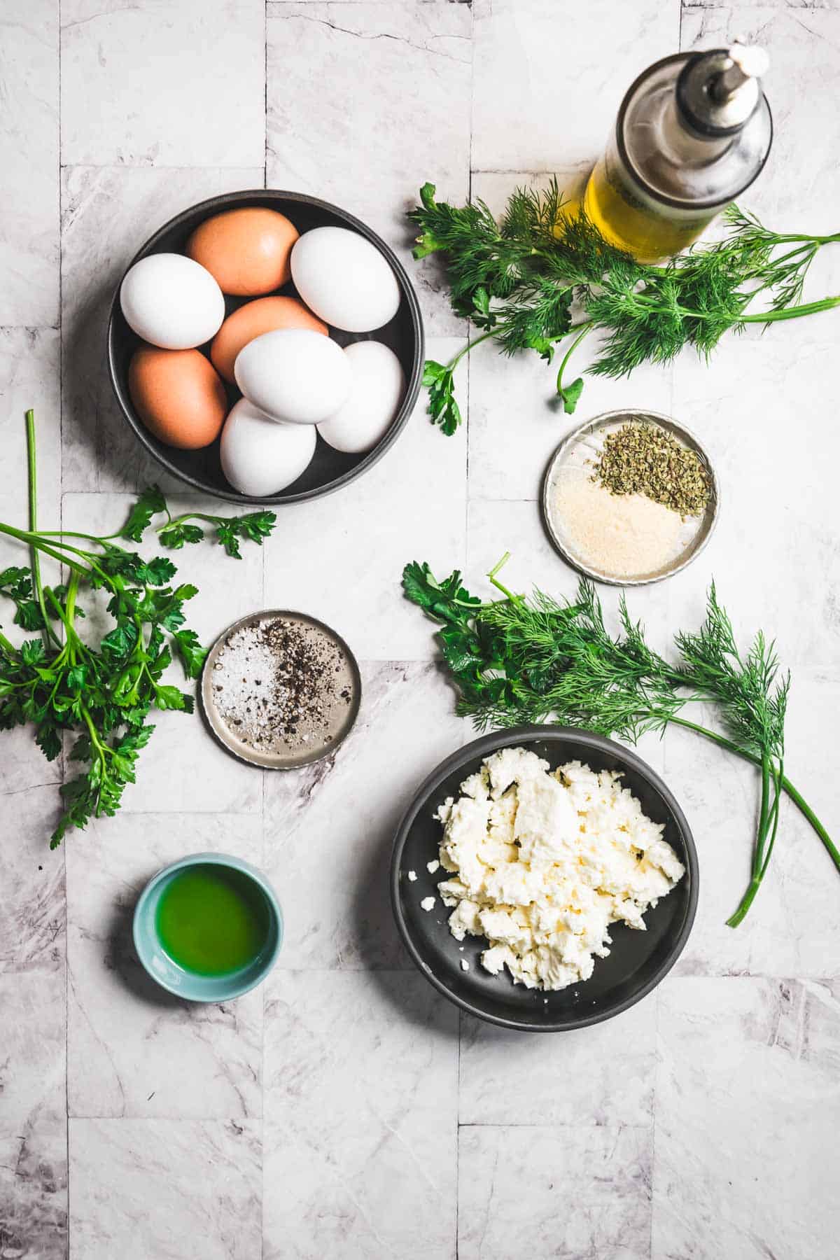 A bowl of eggs, crumbled feta ready for a baked feta and egg salad, fresh herbs, a small bowl of olive oil, and two dishes with seasonings like black pepper rest on a marble surface. A bottle of olive oil sits in the top right corner.