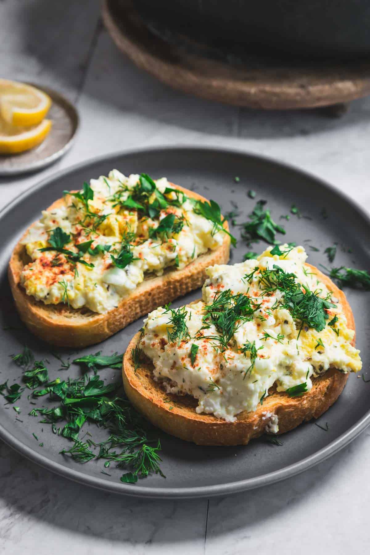 Two slices of bread topped with scrambled eggs, baked feta, herbs, and a sprinkle of paprika are on a gray plate. Fresh dill and parsley are scattered on and around the toast. A lemon slice is visible in the background on a small plate.