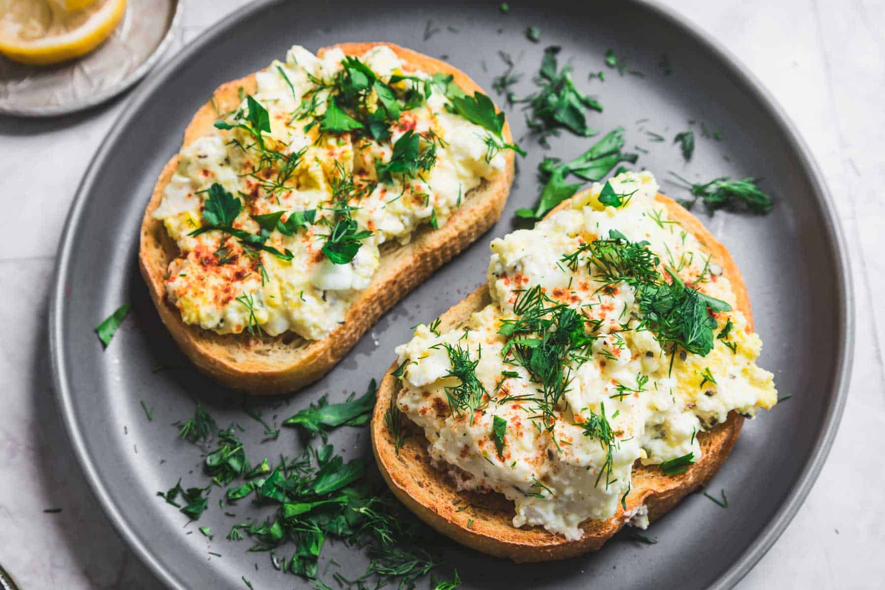 Two slices of bread topped with a savory baked feta and egg salad, garnished with fresh herbs like dill and parsley. The bread rests on a round, gray plate, with scattered herbs along the rim. A lemon wedge peeks from the top-left corner, adding a zesty touch.