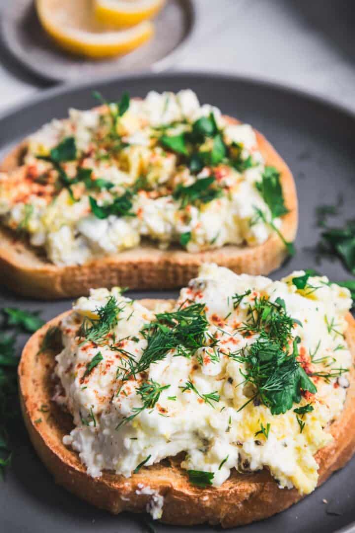 Two slices of toasted bread topped with scrambled eggs, enhanced by hints of dill and paprika, reminiscent of a baked feta and egg salad. The toast is served on a dark plate, with a hint of lemon peeking through in the background.