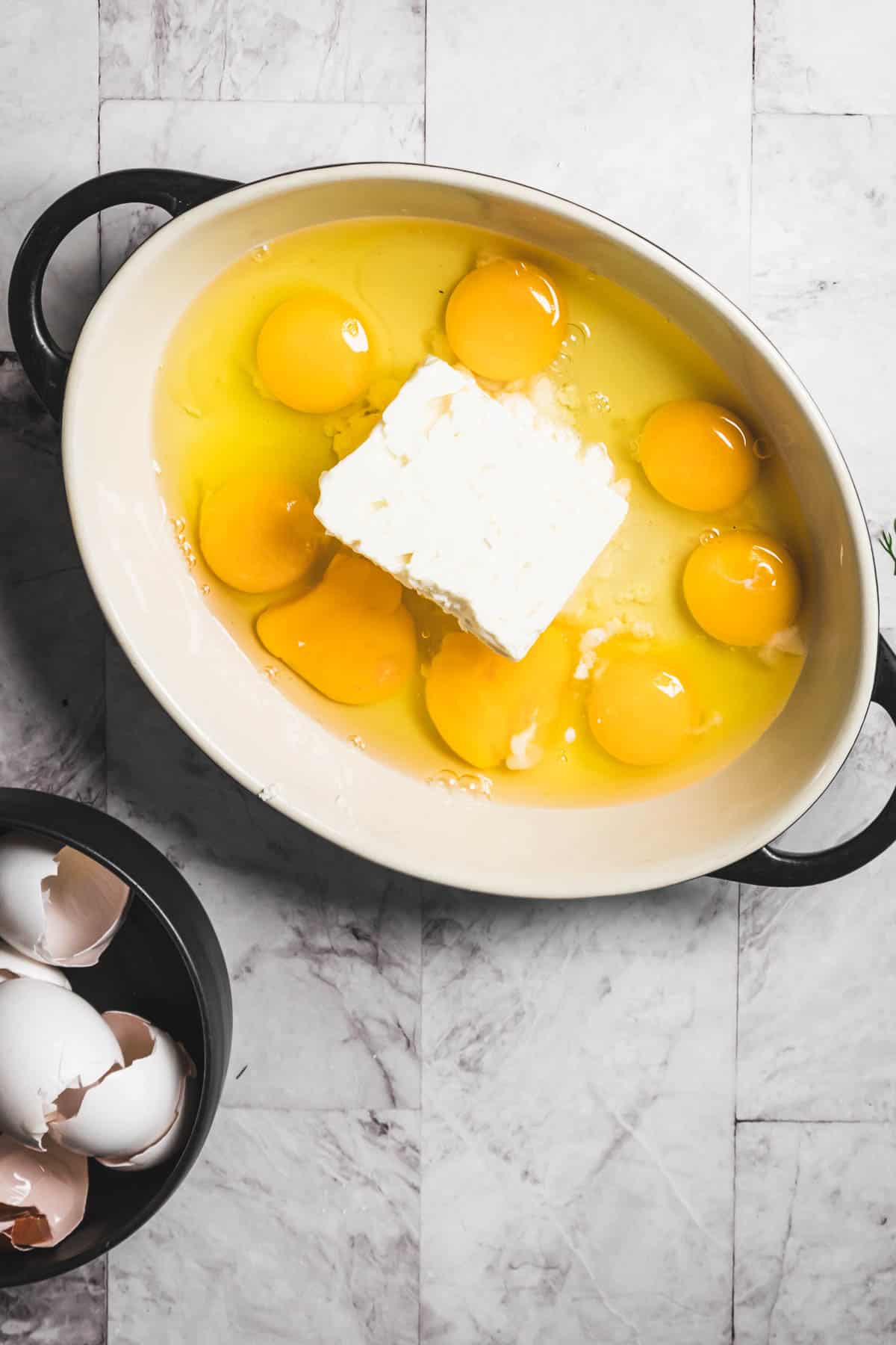 A baking dish on the marble countertop cradles six raw eggs and a block of cheese, ready to transform into a delicious Baked Feta and Egg Salad. Nearby, a small bowl collects the discarded eggshells.