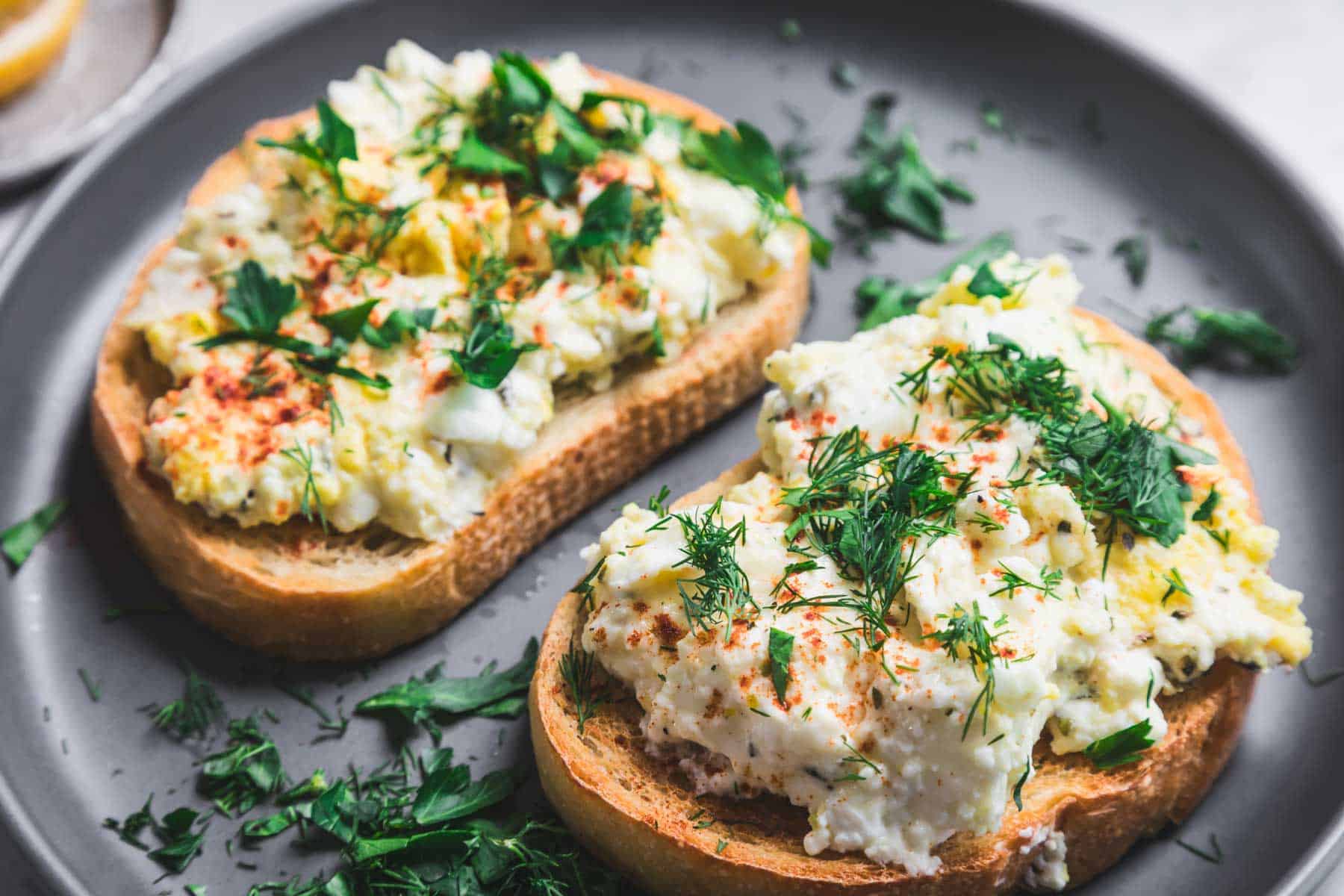 Two slices of toasted bread are topped with a delightful mix of scrambled eggs, baked feta, and garnished with chopped dill and paprika on a gray plate. Additional dill is scattered around the plate.