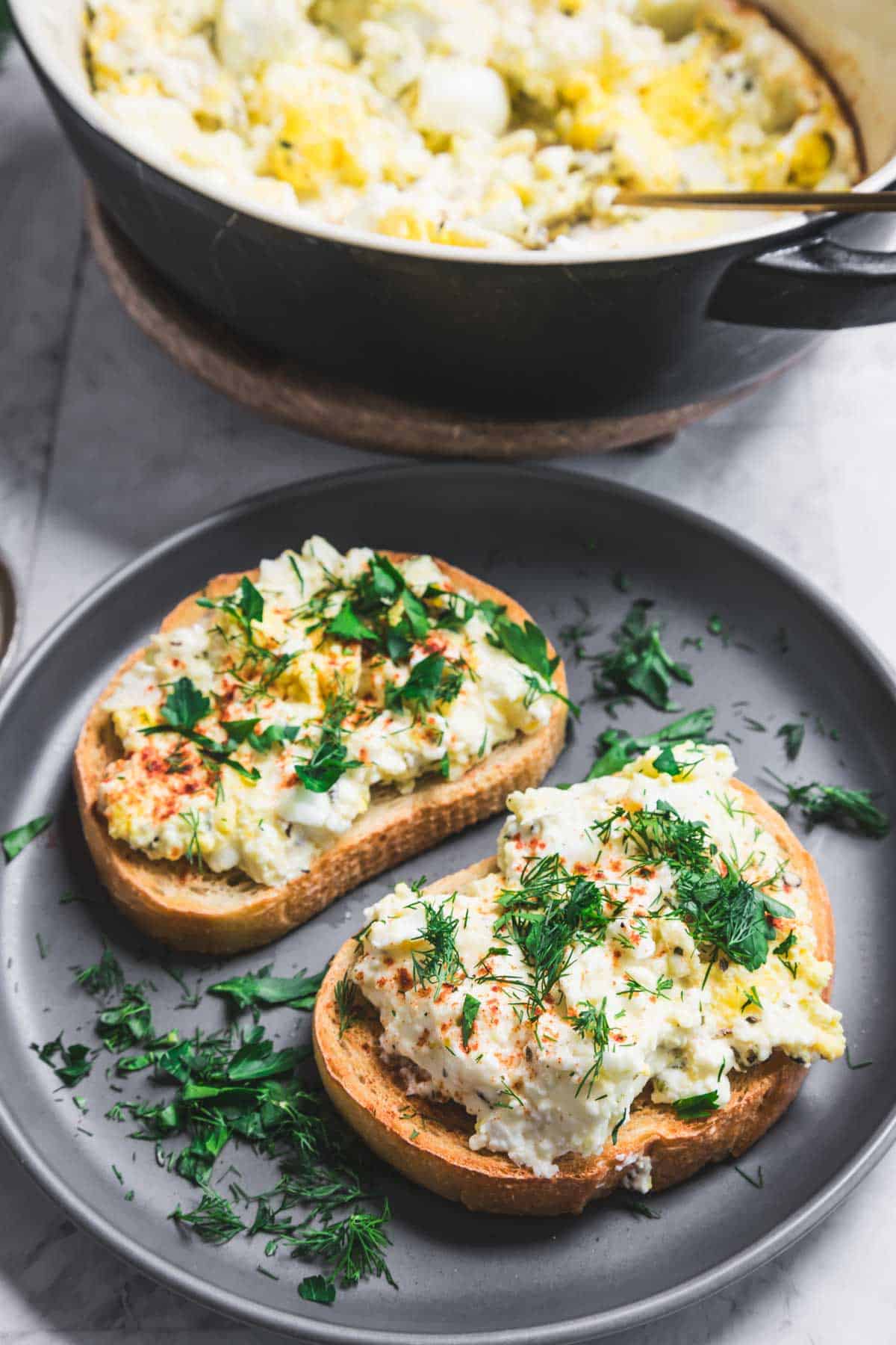 Two slices of toast topped with creamy scrambled eggs and garnished with herbs are on a gray plate, reminiscent of a baked feta and egg salad. A pot containing more scrambled eggs is in the background. Fresh herbs are scattered on the plate and table surface.