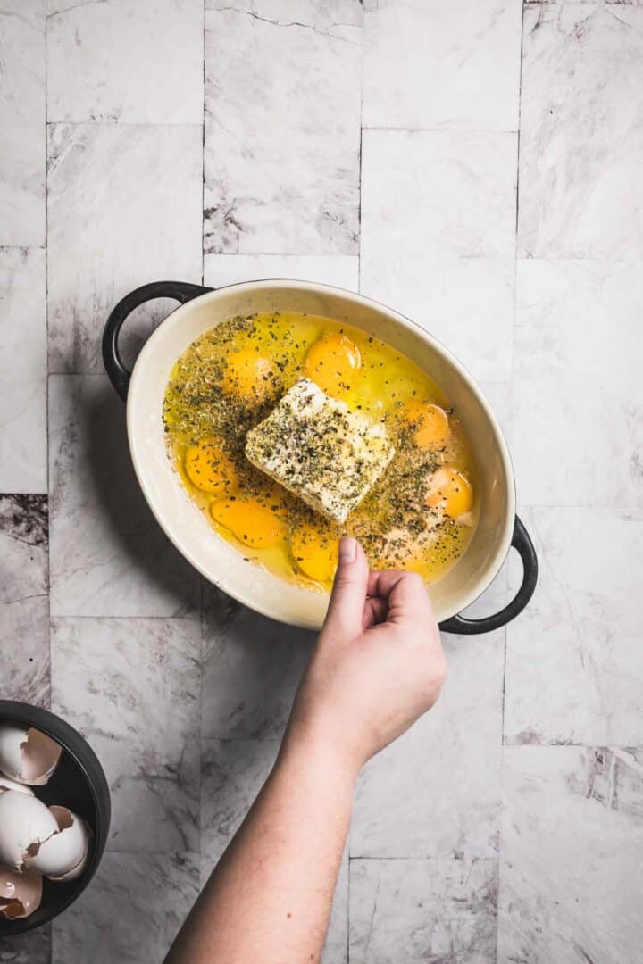 A hand is adding seasoning to a white oval dish of baked feta and eggs with a block of butter. The dish rests on a marble surface, exuding an inviting aroma. Nearby, a small bowl holds cracked eggshells, hinting at the salad's fresh preparation.