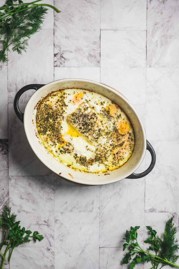 A white oval baking dish with handles contains a savory baked feta and egg salad topped with herbs. The dish rests on a tiled surface, accompanied by sprigs of fresh green herbs strategically placed around it.