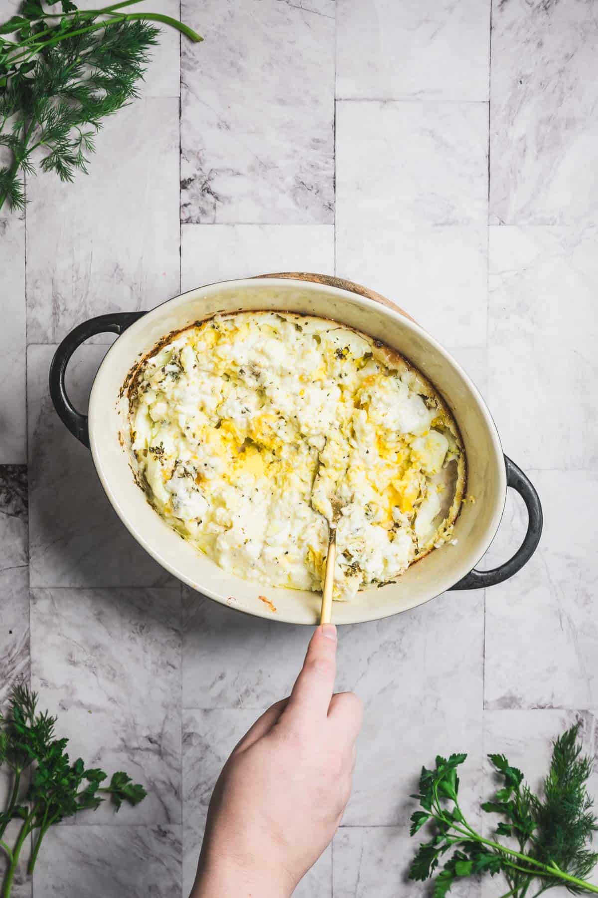 A hand holds a spoon, serving from a baked dish of mashed potatoes topped with melted cheese in a white oval casserole dish. The dish, reminiscent of comfort food like baked feta and egg salad, is set on a gray tiled surface, with sprigs of parsley around it.