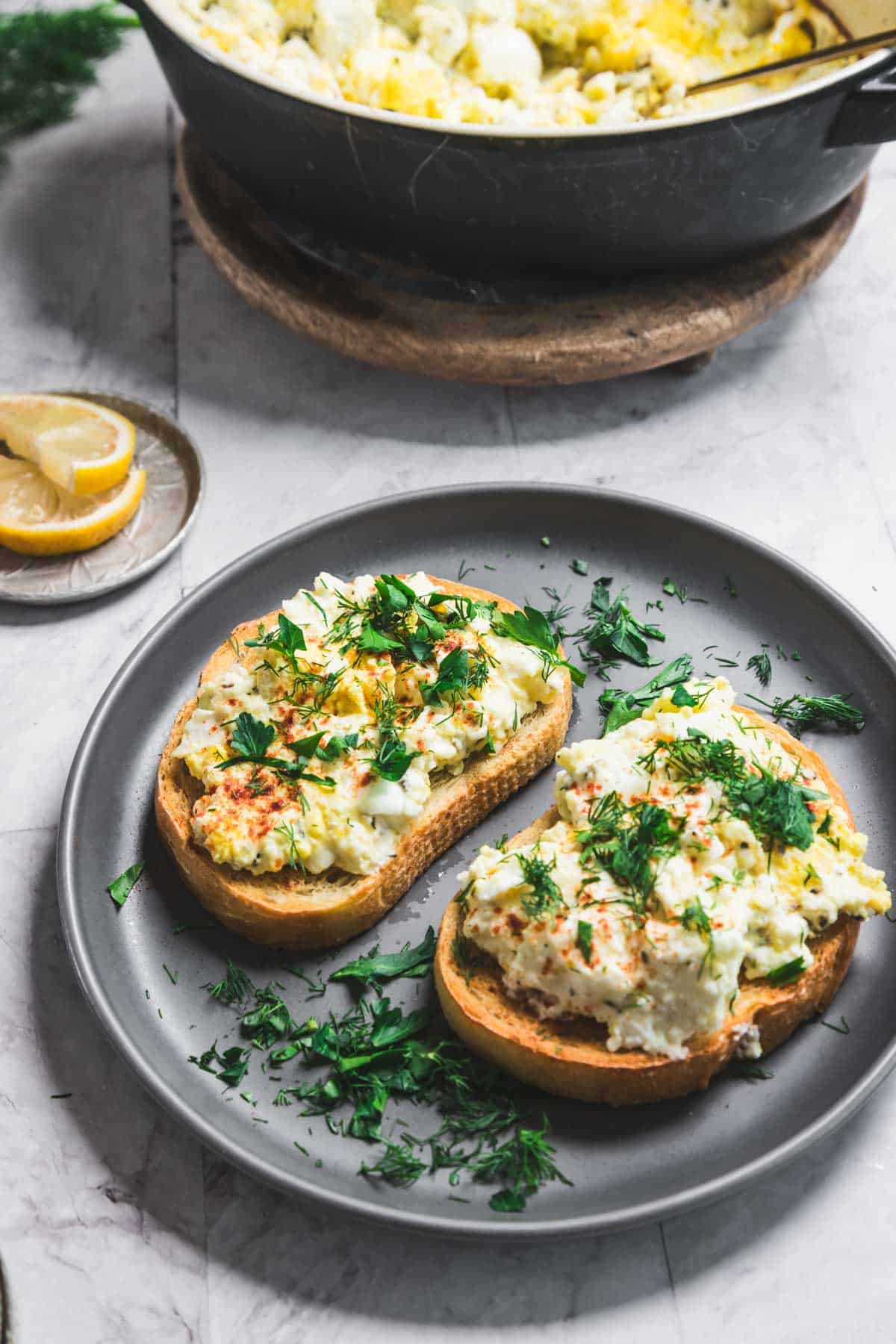 A plate with two slices of toast topped with creamy scrambled eggs, enhanced by a hint of baked feta, garnished with fresh herbs and a sprinkle of paprika. A lemon slice and additional herbs are on the side. A pan of scrambled eggs is partially visible in the background.