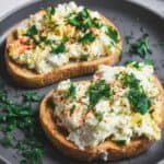 Two slices of toasted bread topped with a creamy baked feta and egg salad, garnished with fresh dill and parsley, served on a gray plate.