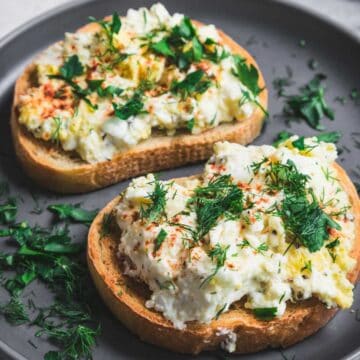 Two slices of toasted bread topped with a creamy baked feta and egg salad, garnished with fresh dill and parsley, served on a gray plate.