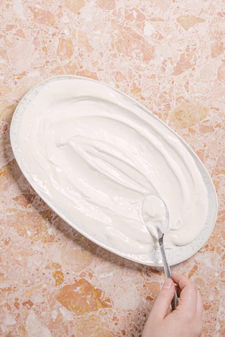 A hand is using a spoon to spread a thick, creamy white substance evenly across an oval plate. The plate is placed on a marbled pink and beige surface.