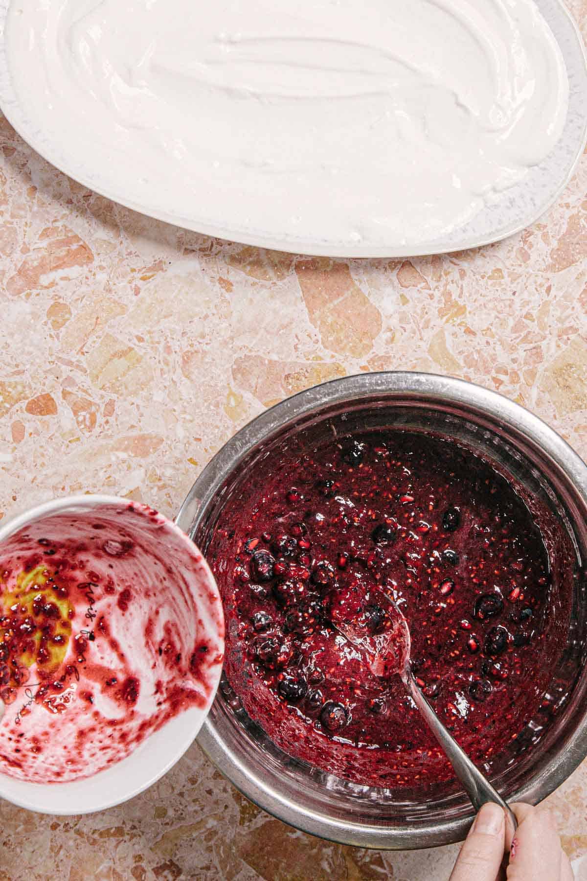 A bowl with a red berry mixture is on a marble surface, next to an empty smaller bowl with berry residue. A hand holds a spoon in the large bowl. A white dish with a creamy white layer is partially visible above.