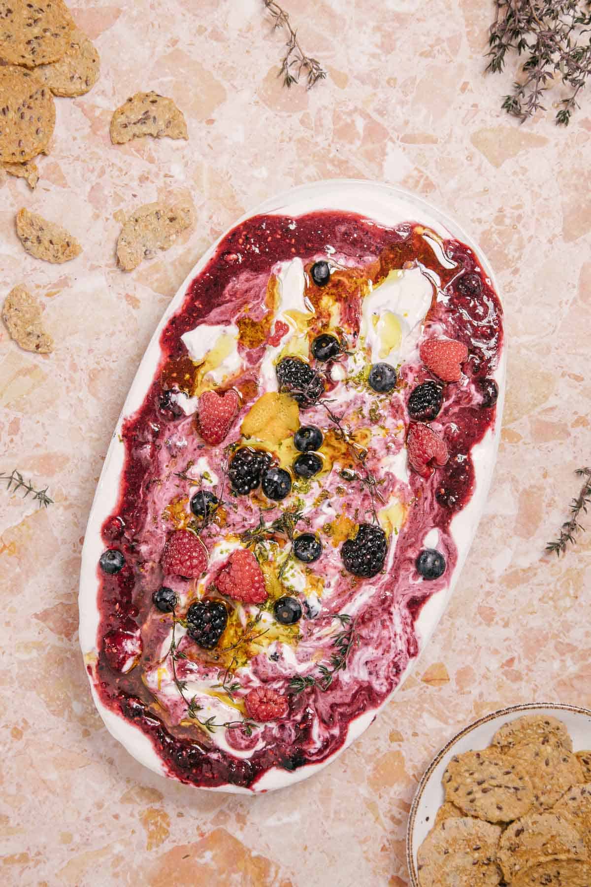 Oval platter with whipped goat cheese topped with mixed berry compote, fresh raspberries, blackberries, blueberries, and thin lemon slices. Surrounded by scattered herbs and crackers on a marbled surface.