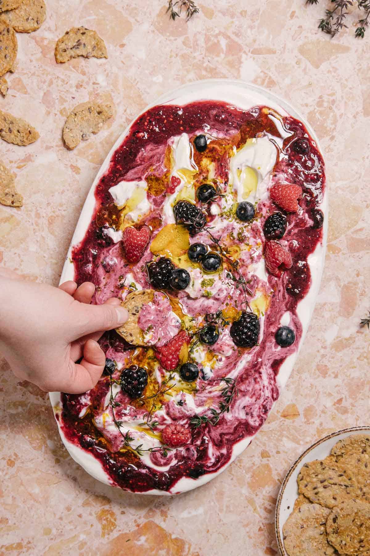 A hand dips a cracker into a spread of mixed berries on creamy yogurt or cheese, garnished with honey and thyme, on an oval plate. The dish rests on a textured marble surface, surrounded by more crackers and scattered herbs.